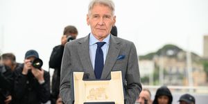harrison ford, wearing a gray suit and blue tie, holds the palm d or award on a stage while photographer behind him take pictures