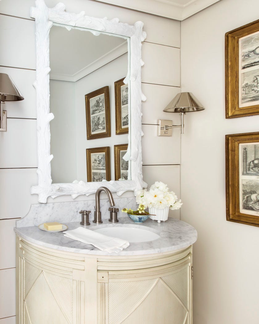 powder bathroom with white wood framed mirror, white marble vanity top with small vase of white flowers, metal wall lamps, and framed prints on far wall