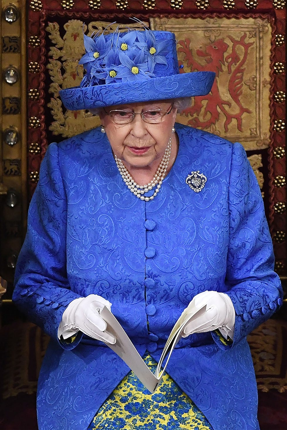 Queen Elizabeth II during the 2017 Queen's Speech. She wears a blue hat with blue and yellow flowers, and a blue and yellow floral dress.