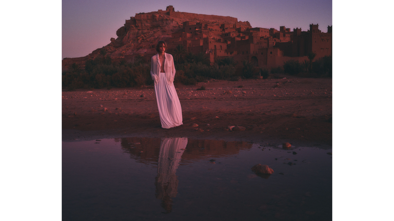 silhouette of a person wearing a long white dress standing near a reflective pool with a mountainous fortress in the background