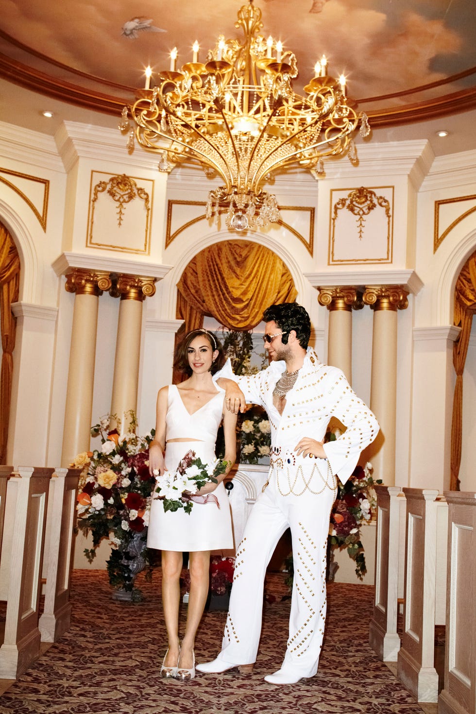 a couple dressed in formal outfits stands in a richly decorated interior