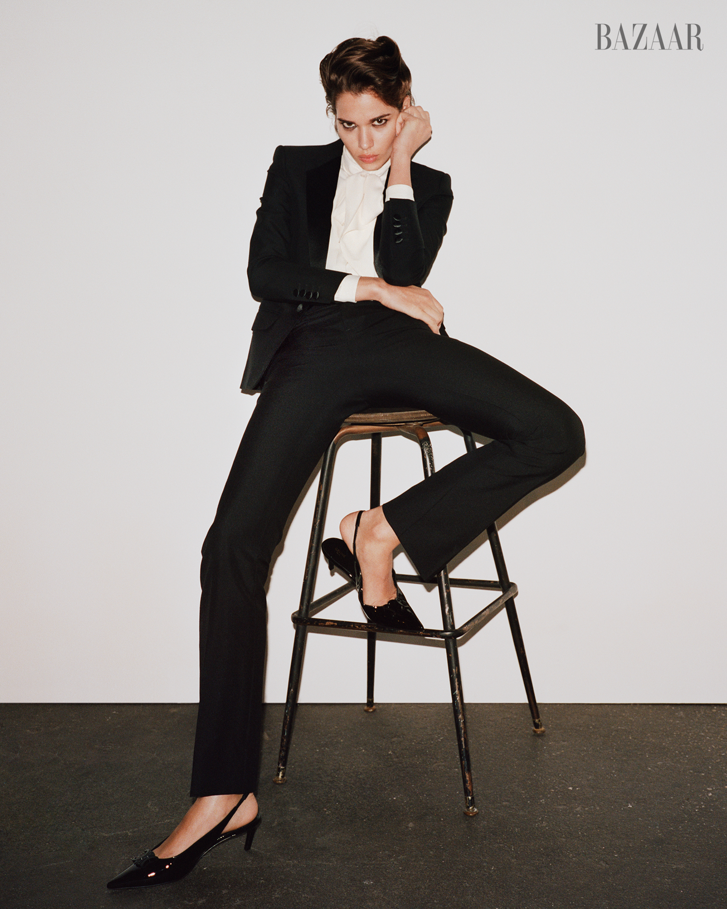 a model poses in a stylish black suit and heels seated on a stool