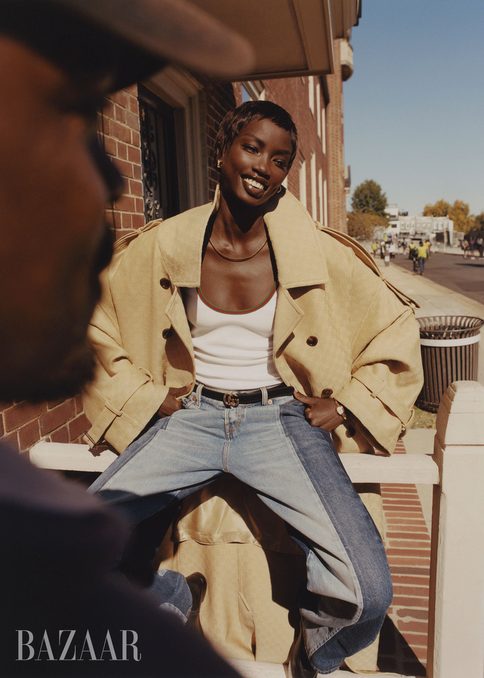 fashion model wearing a beige coat and blue jeans seated in an urban setting