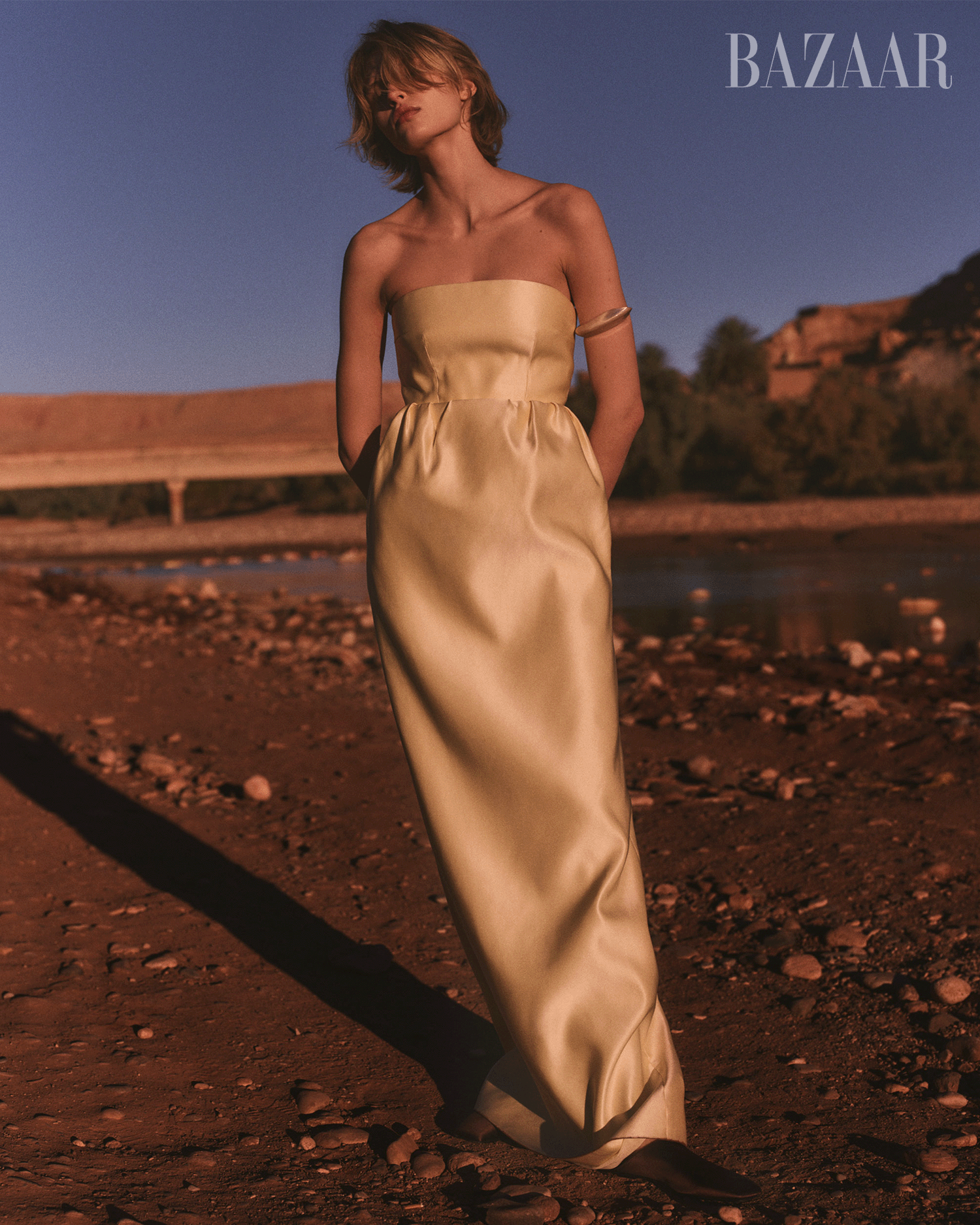 a model wearing a yellow strapless gown standing on a rocky riverbank at sunset