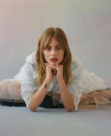 samara sits on floor with her chin resting on top of clasped hands she wears a white button up and a beige tulle skirt
