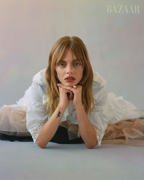 samara sits on floor with her chin resting on top of clasped hands she wears a white button up and a beige tulle skirt