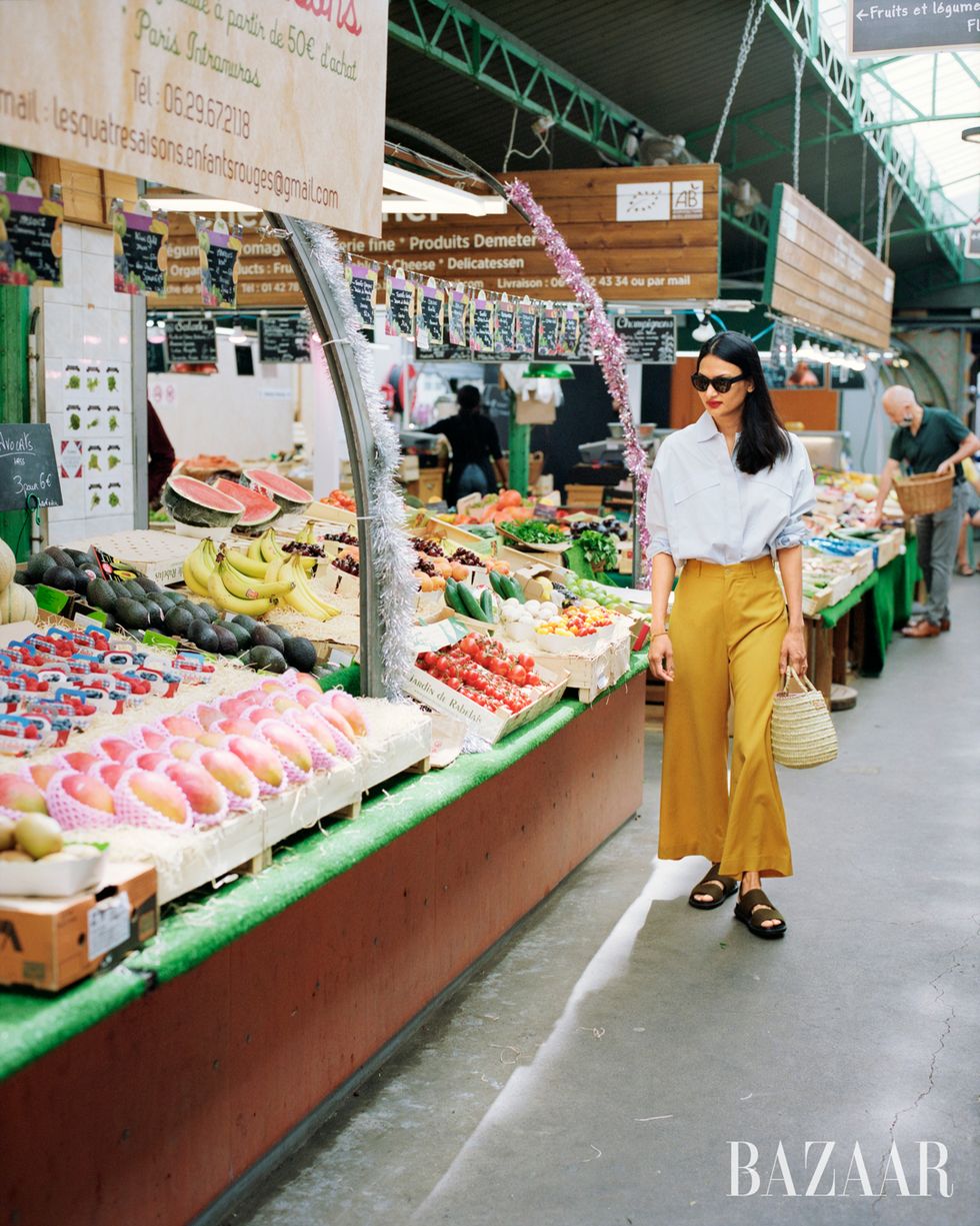 woman in yellow pants white shirt and tan woven basket bag approaching fruit at supermarket