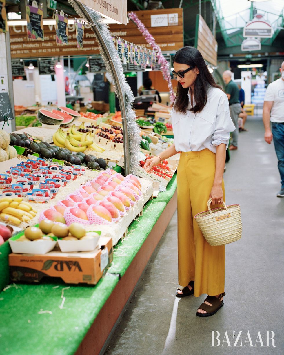 woman in yellow pants white shirt and woven basket bag looking at fruit at market