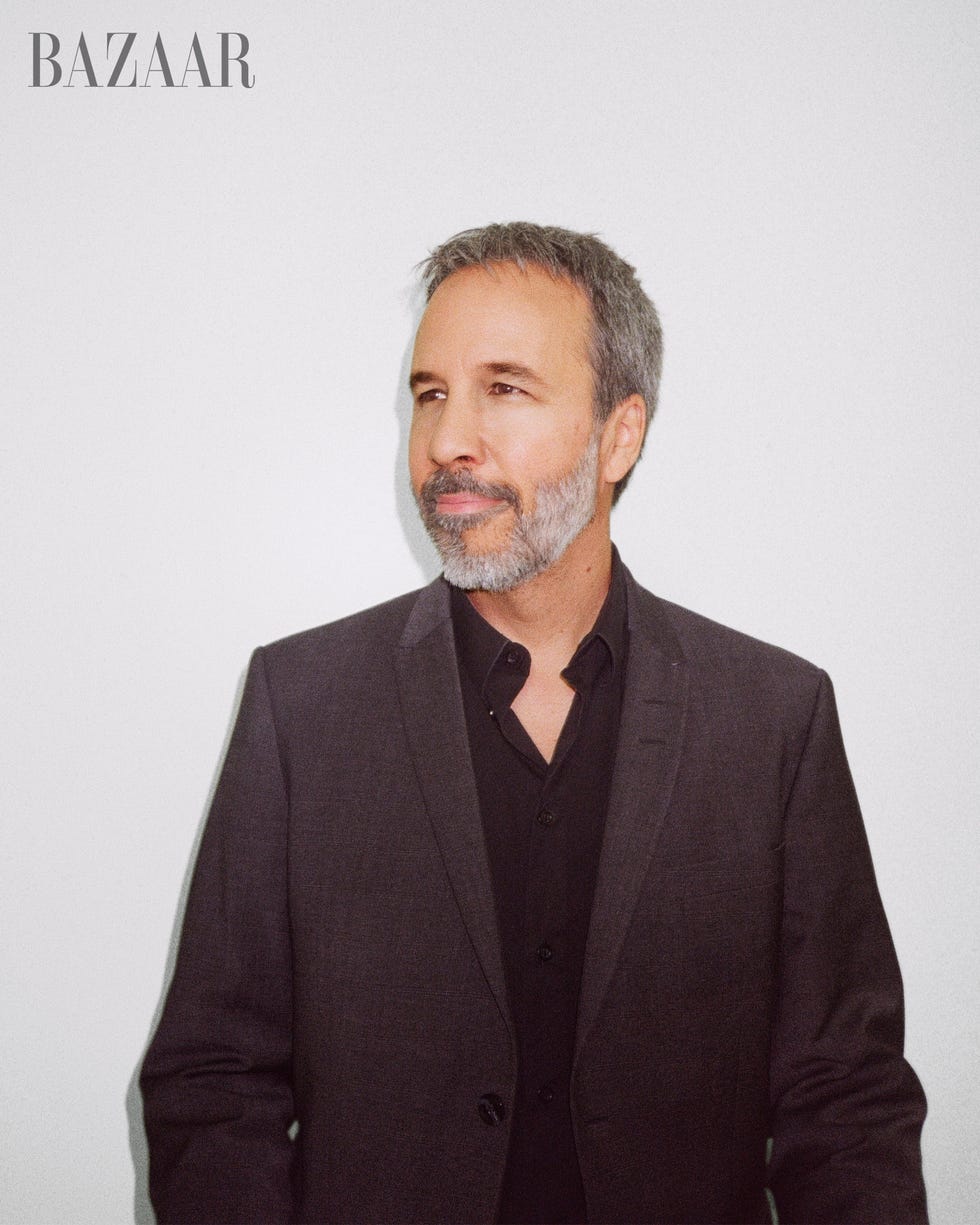 denis sits against white wall wearing a dark gray blazer and black pants and a black button up looking off camera