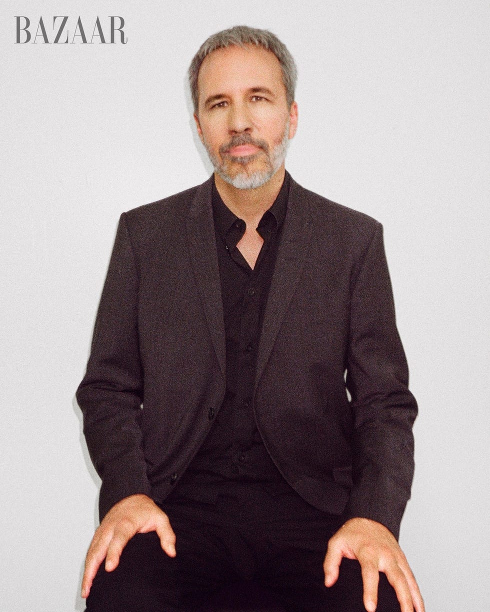 denis sits against white wall looking directly at camera, wearing a gray blazer and black slacks and a black button up