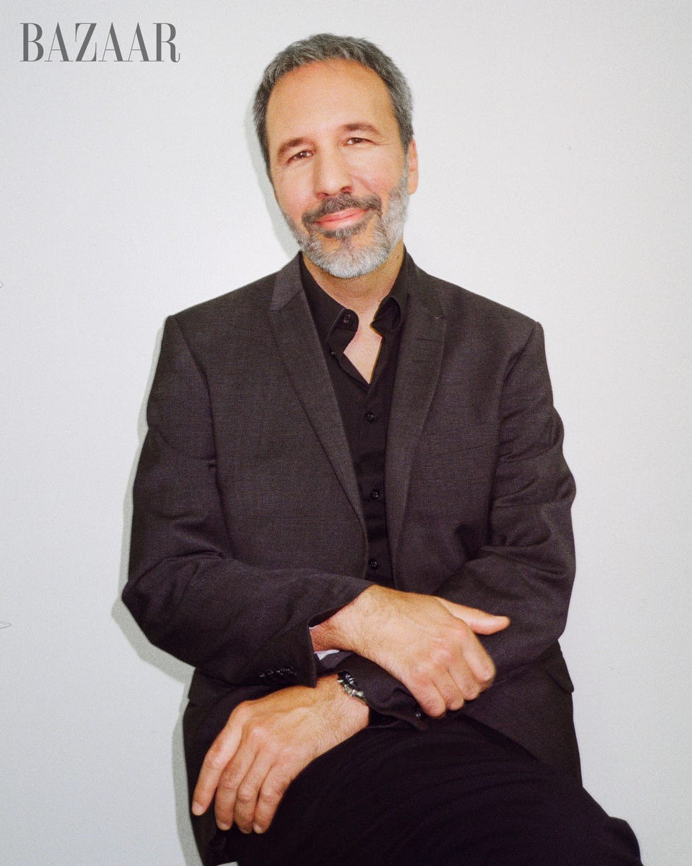 denis sits against white wall looking directly at camera, with his arms crossed in front of him, wearing a gray blazer and black slacks and a black button up