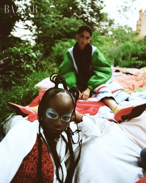 two women laying on grass, bottom in light blue eyeshadow and red patterned shirt, top in green jacket