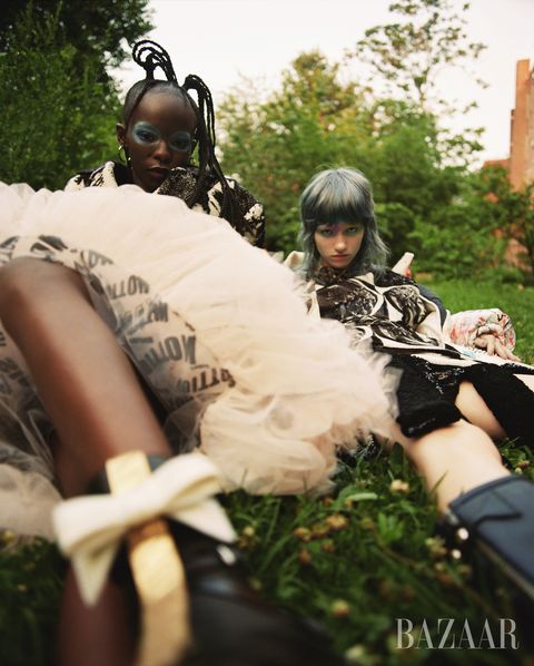 two models sit on grass with dramatic eye makeup, one wears a tulle skirt and the other a dress with boots