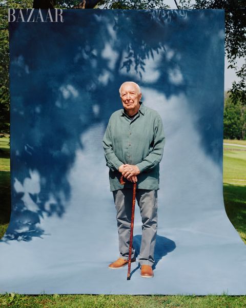 jasper stands in front of a blue background set up in a field, he poses in a gray green blue button up, gray slacks, brown shoes, and holding a brown walking cane