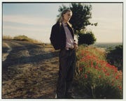 model stands on a dirt path lined by red flowers and overlooking a view wearing a lavender button up, a sweater, and trousers