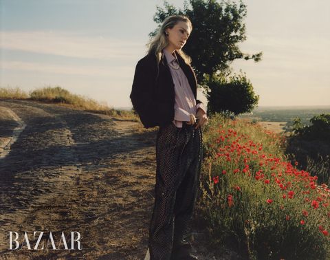 blonde woman in lavender blouse, chevron black trousers, black coat next to red flowers