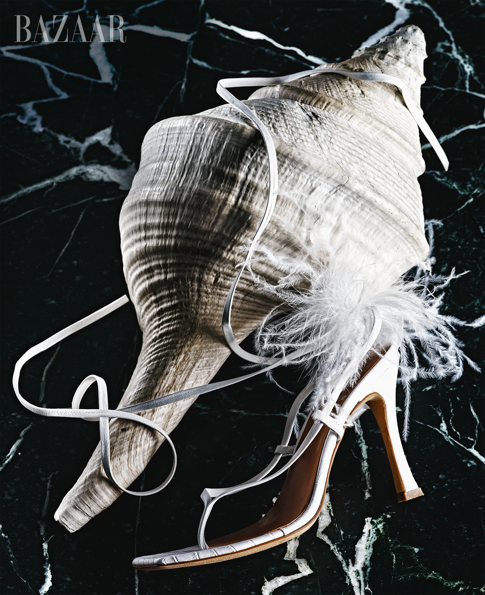 a strappy white sandal designed by brother vellies for bloomingdale's 150th anniversary collection, with one long white strap wrapped around a large conch shell, shot on a dark green marble background