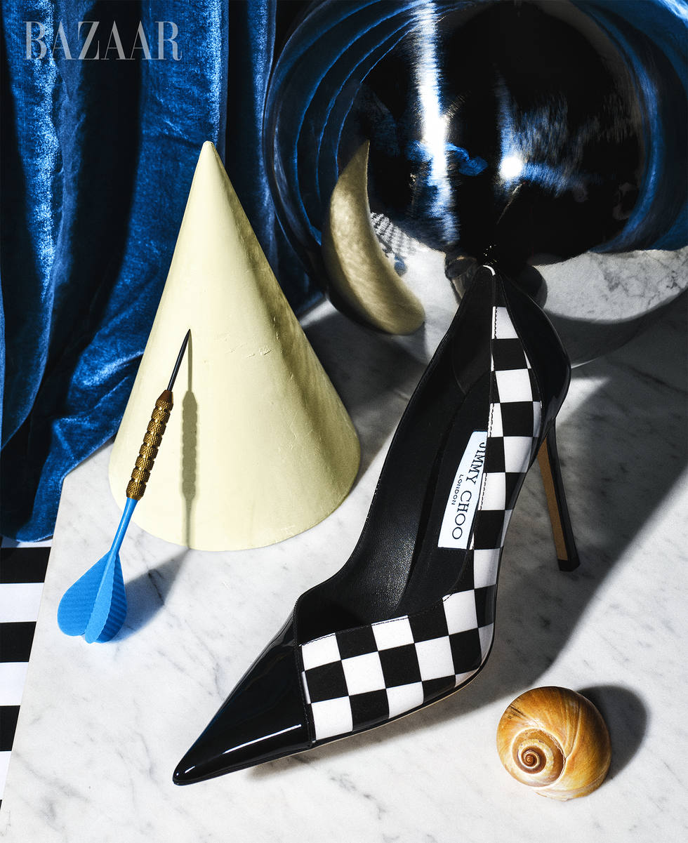 a black and white checkered heel designed by jimmy choo for bloomingdale's 150th anniversary collection shot on a surreal background with a seashell, a cone, and a dart on a marble slab in front of a blue velvet curtain