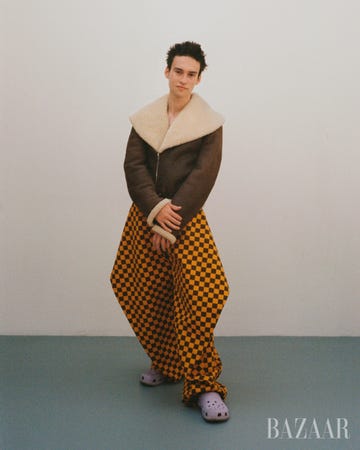 jacob collier posing in brown moto jacket with fur trim yellow and black checkered pants and light purple crocs