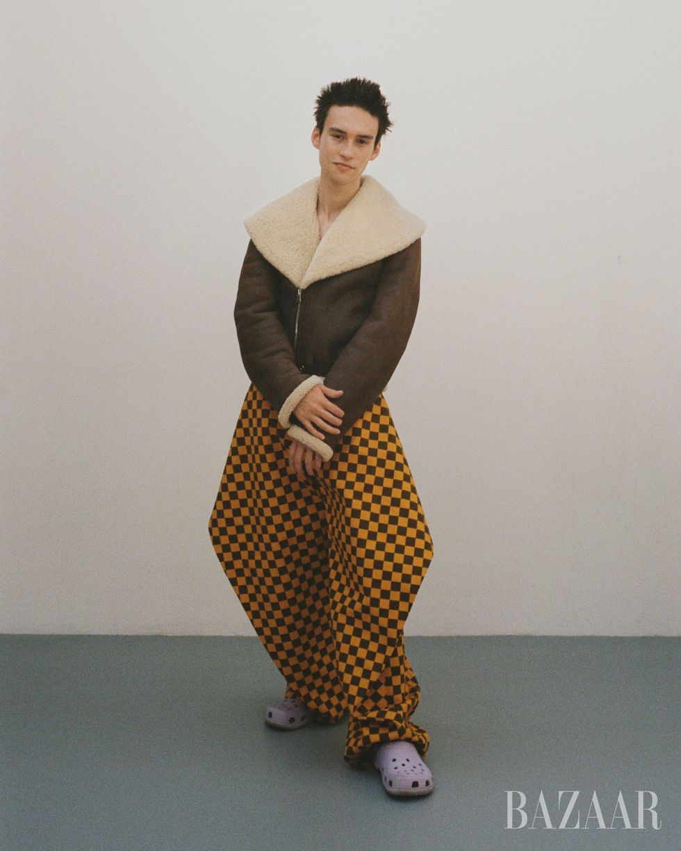 jacob collier posing in brown moto jacket with fur trim yellow and black checkered pants and light purple crocs
