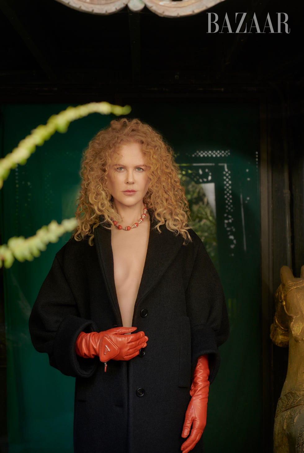 nicole stands in front of a reflective green wall wearing a black undone trench coat and red leather gloves