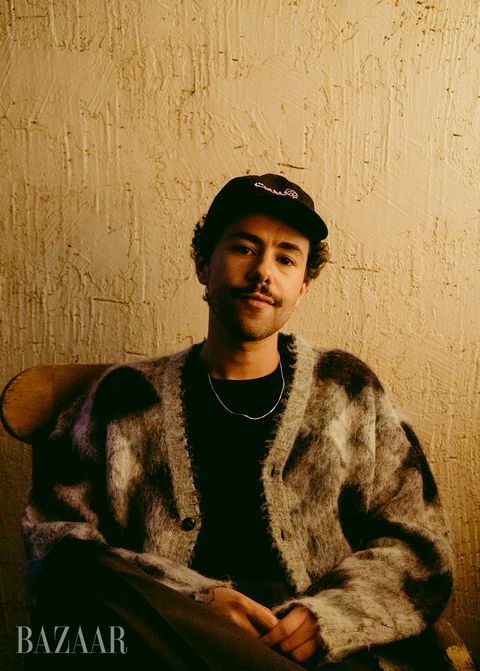 a portrait of ramy youssef wearing a black and gray plaid cardigan, a black tee, a thin gold chain, and a black baseball hat