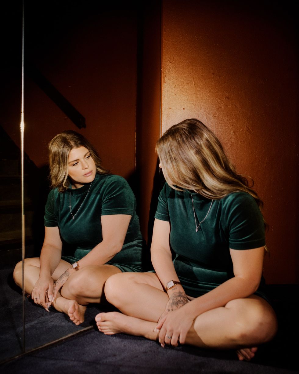 artist yebba sits cross legged looking at herself in a mirror wearing a green velour top and a gold necklace