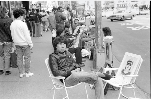 a young ai weiwei sits on a folding chair wearing a dark leather jacket and trousers in 1987