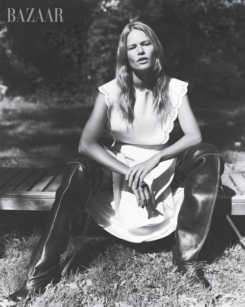 black and white image of model anna ewers sitting on wooden platform in white dress and black boots