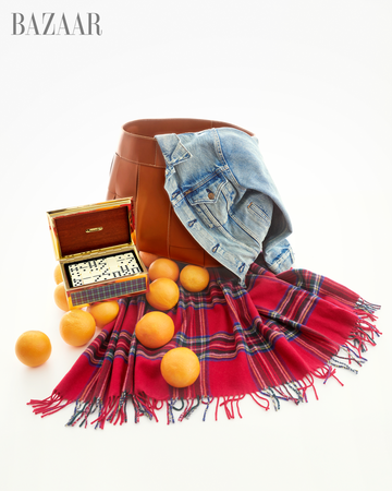 A stylish flat lay featuring a brown bag a denim jacket a vibrant red plaid scarf scattered oranges and a decorative box