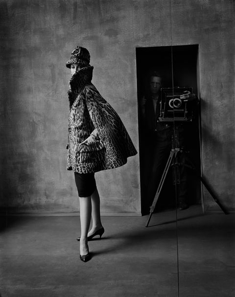 a model in a leopard cape and matching hat, a pencil skirt and kitten heels stands in front of a door, while photographer Melvin Sokolsky stands with his camera in the background