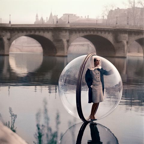 a model wearing a checkered suit and white headpiece stands in a large bubble in front of a bridge in paris