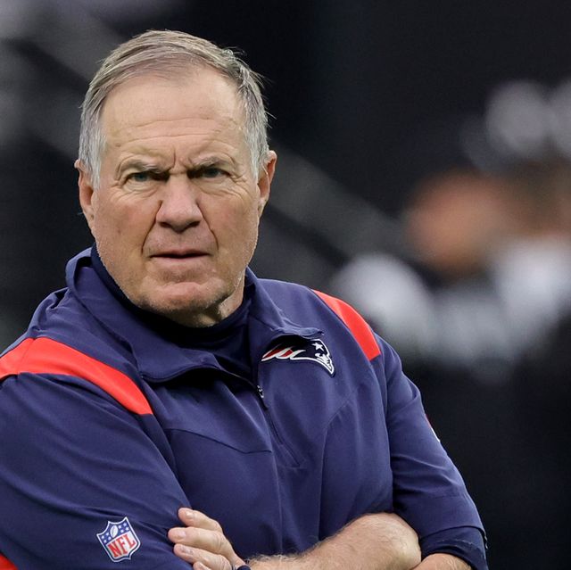 bill belichick looks angrily toward the camera with his arms crossed, he wears a blue new england patriots vest and matching shirt with an nfl logo on the right sleeve