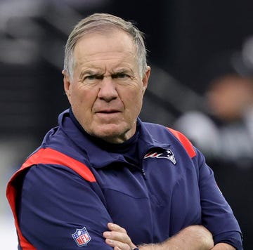 bill belichick looks angrily toward the camera with his arms crossed, he wears a blue new england patriots vest and matching shirt with an nfl logo on the right sleeve