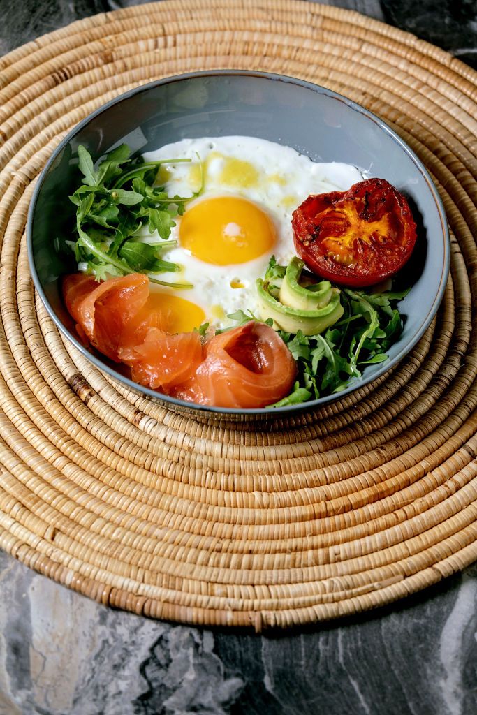 healthy breakfast bowl with fried eggs salmon avocado grilled tomato and salad serving with bread on straw napkin over dark marble background