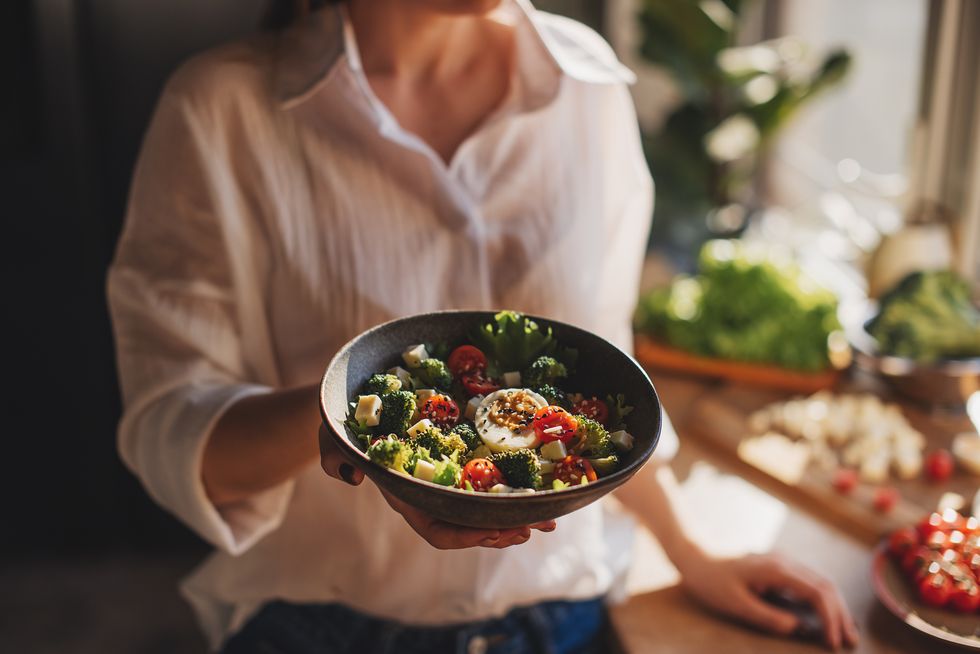 healthy dinner or lunch woman in t shirt and jeans standing and holding vegan superbowl or buddha bowl with hummus, vegetable, salad, beans, couscous and avocado and smoothie in hands, square crop