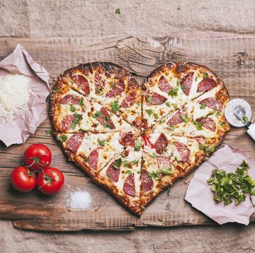 sliced heart shaped pizza with toppings on a wood cutting booard with tomatoes, basil, shredded cheese and pizza cutter