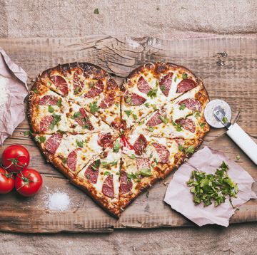 sliced heart shaped pizza with toppings on a wood cutting booard with tomatoes, basil, shredded cheese and pizza cutter