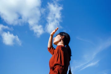 low angle portrait of young asian woman shielding eyes with her hand, standing against beautiful clear blue sky and sunlight. freedom in nature. connection with nature