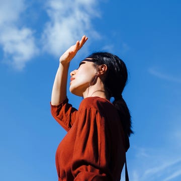 What does the heat do to your body and brain low angle portrait of young asian woman shielding eyes with her hand, standing against beautiful clear blue sky and sunlight. freedom in nature. connection with nature