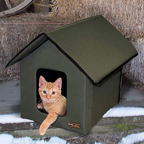 small orange cat in a heated green cat house on a rustic porch with haybells a wooden barrel and a wooden wheel in the background snow is on the ground
