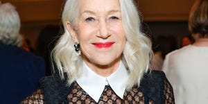 helen mirren smiles at the camera, she wears a black dress with a sheer top and matching white collar and cuffs, she stands inside a crowded room with people behind her