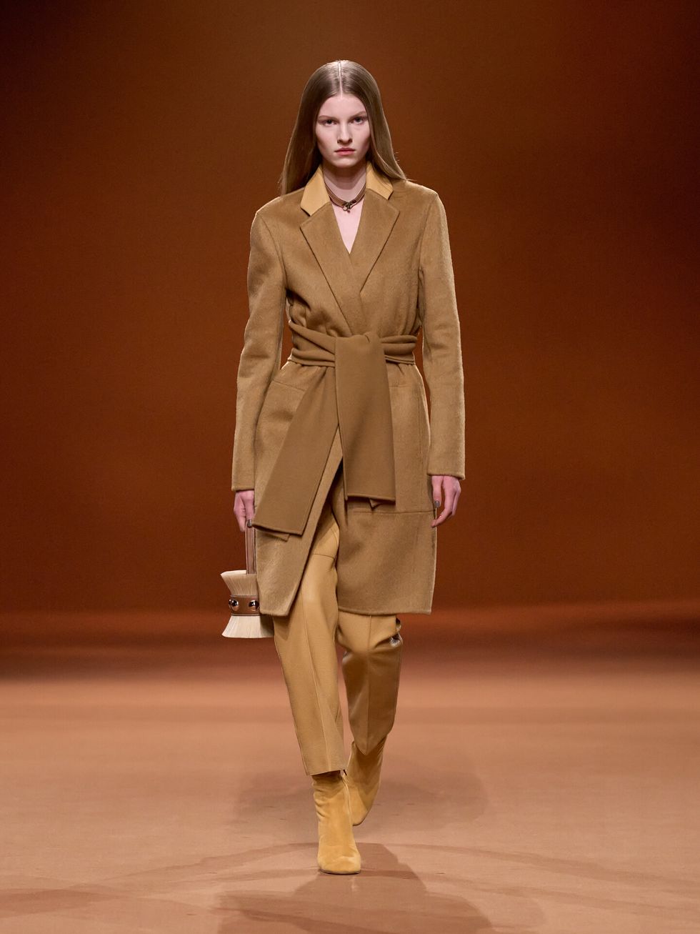 a brunette model walks the hermes fall 2023 runway wearing a coat, trousers, and boots in a sort of matte gold color