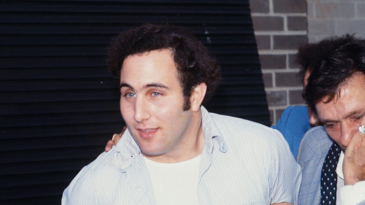 a man walks outside as he looks forward to the left, he wears a collared shirt over a white tshirt, men walk on the right