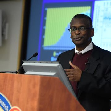 lonnie johnson stands behind a wooden lectern and speaks into a microphone, he wears a black suit jacket, maroon sweater, white collared shirt and tie, behind him is a screen projection showing two charts