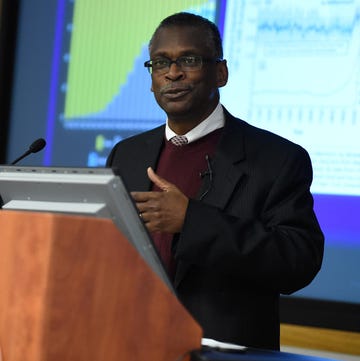 lonnie johnson stands behind a wooden lectern and speaks into a microphone, he wears a black suit jacket, maroon sweater, white collared shirt and tie, behind him is a screen projection showing two charts