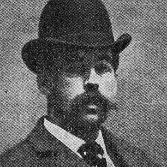 hh holmes looks at the camera, he wears a bowler hat, jacket, collared shirt and tie and has a mustache