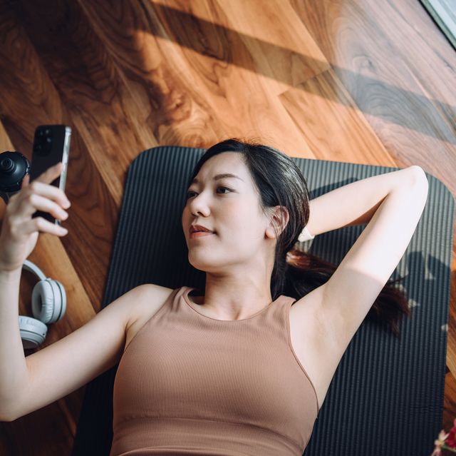 high angle shot of fitness young asian sports woman relaxing after working out, lying on yoga mat and using her smartphone against sunlight in the morning, reusable water bottle and headphones by her side health, fitness and wellness concept