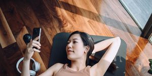 high angle shot of fitness young asian sports woman relaxing after working out, lying on yoga mat and using her smartphone against sunlight in the morning, reusable water bottle and headphones by her side health, fitness and wellness concept