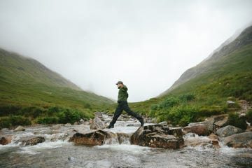 hiker crossing river in mountain valley, glencoe, scotland